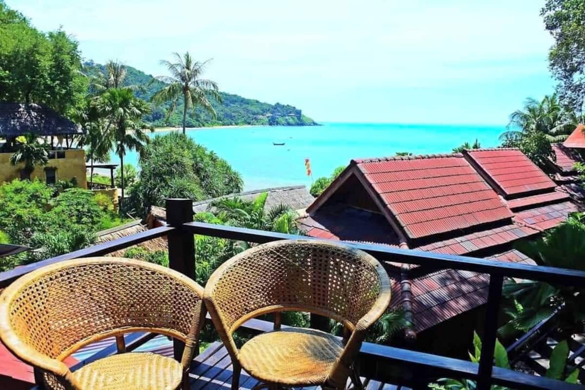 A balcony with wicker chairs overlooking a beach with clear blue water and a small boat in the distance.