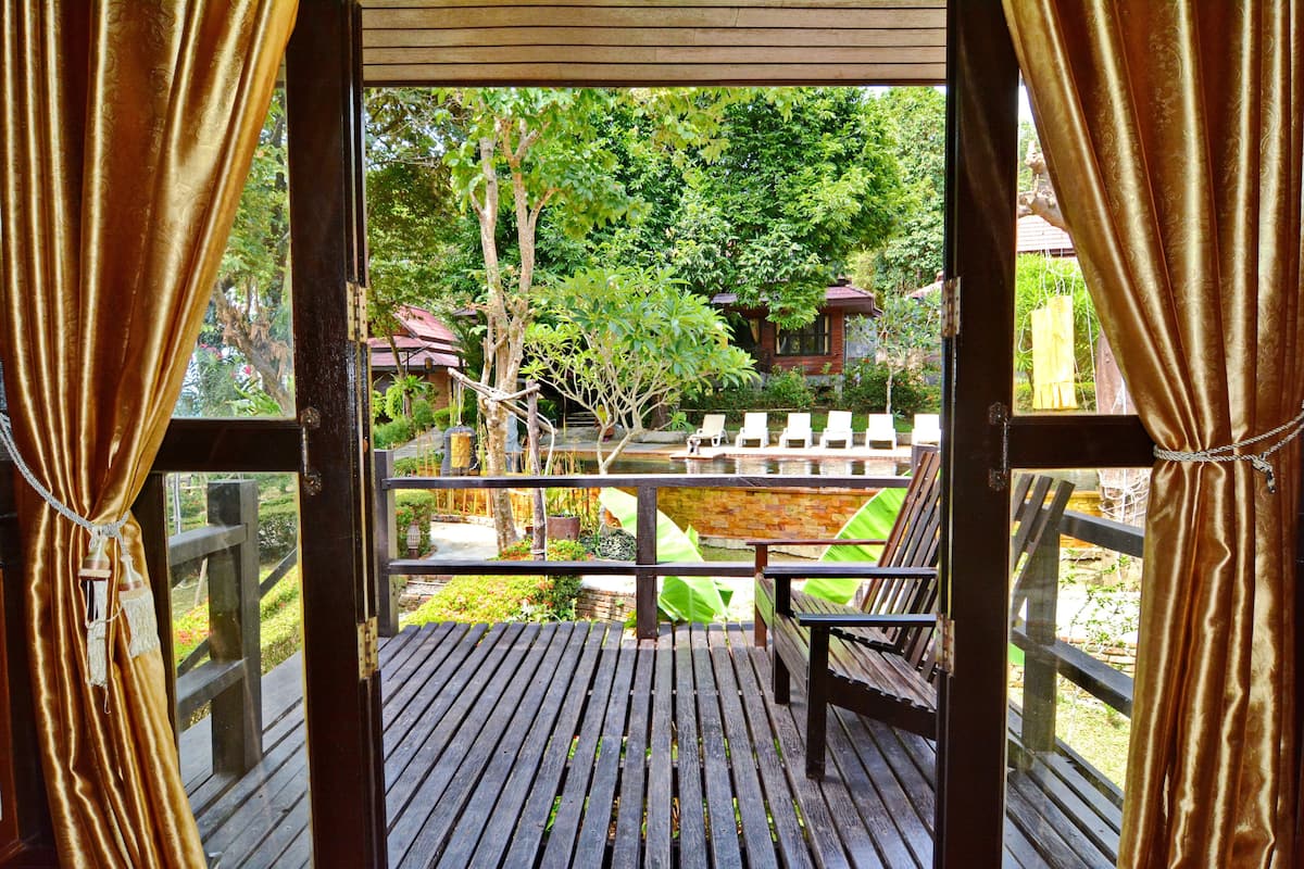 A wooden deck with a bench, a small pond, and a view of a garden with trees and a building in the background.