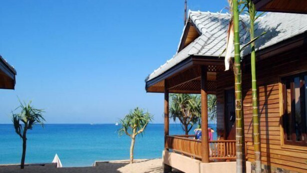 Een houten strandhuis met een overdekte veranda, uitzicht op zee en een palmboom op de voorgrond.
