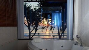 Een badkamer met een badkuip, een raam met zonwering en uitzicht op palmbomen en een zonsondergang.