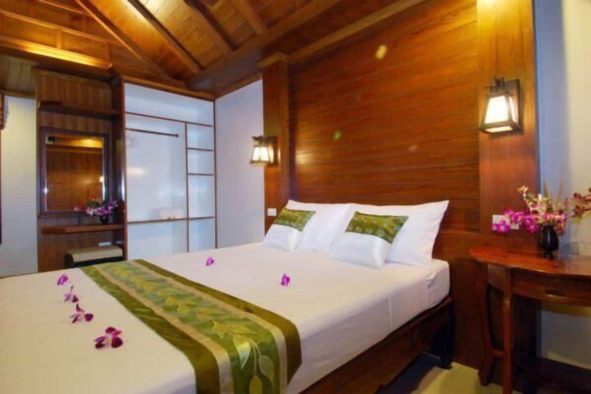 Ein Hotelzimmer mit Holzbalkendecke, einem Bett mit weißen Leinentüchern und grünen Kissen, einem Nachttisch mit Blumenvase und einem Holzschrank.