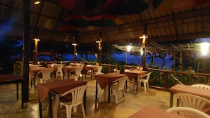 Een buitenterras met tafels en stoelen, een rieten dak en uitzicht op de oceaan bij zonsondergang.