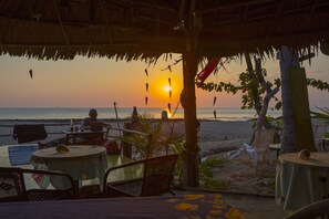 A beachside restaurant with tables and chairs under a thatched roof, overlooking the ocean at sunset.