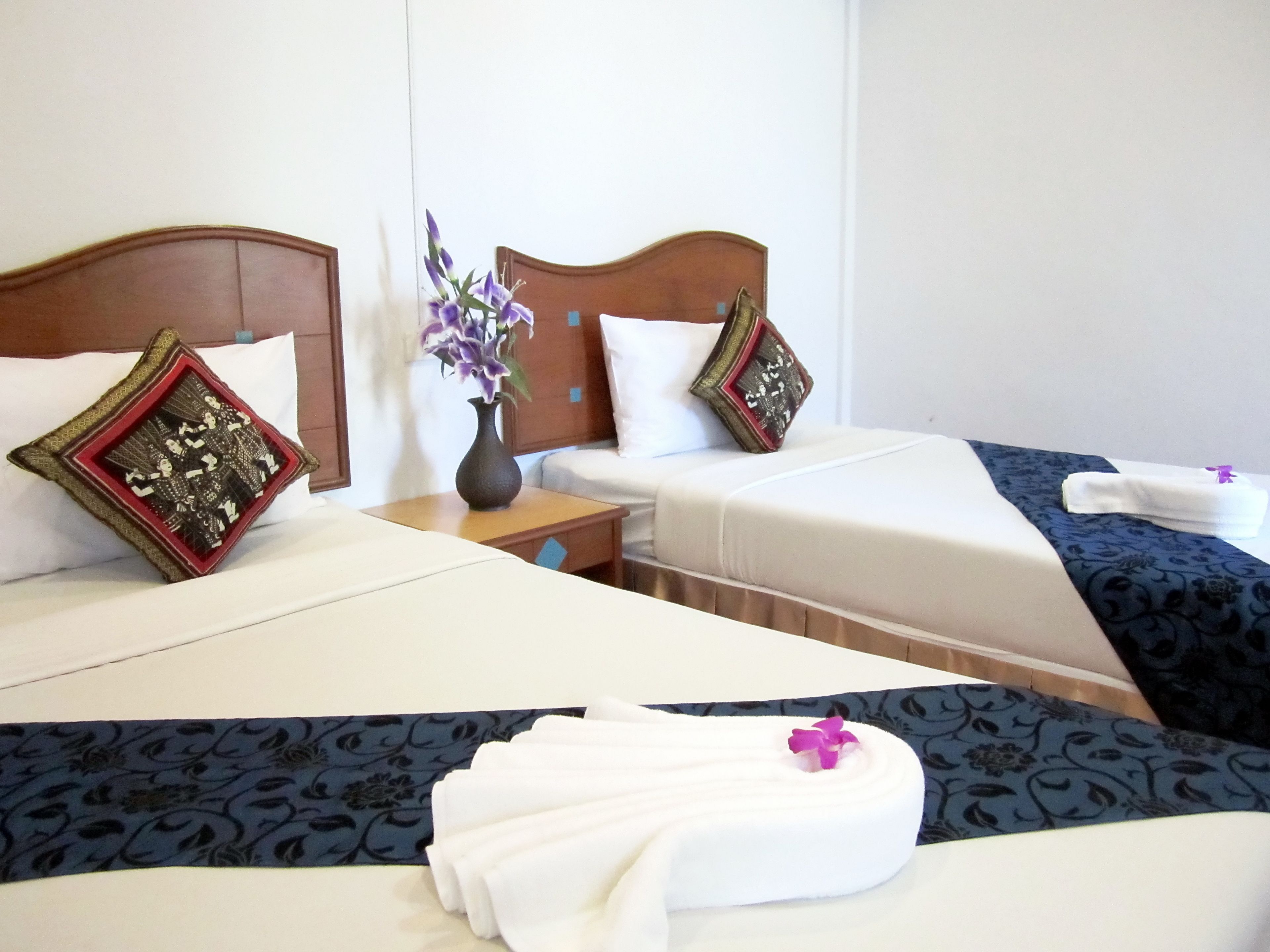 Habitación de hotel con dos camas, cada una con una almohada decorativa, una mesita de noche con un florero y una toalla doblada con cuidado sobre la cama.