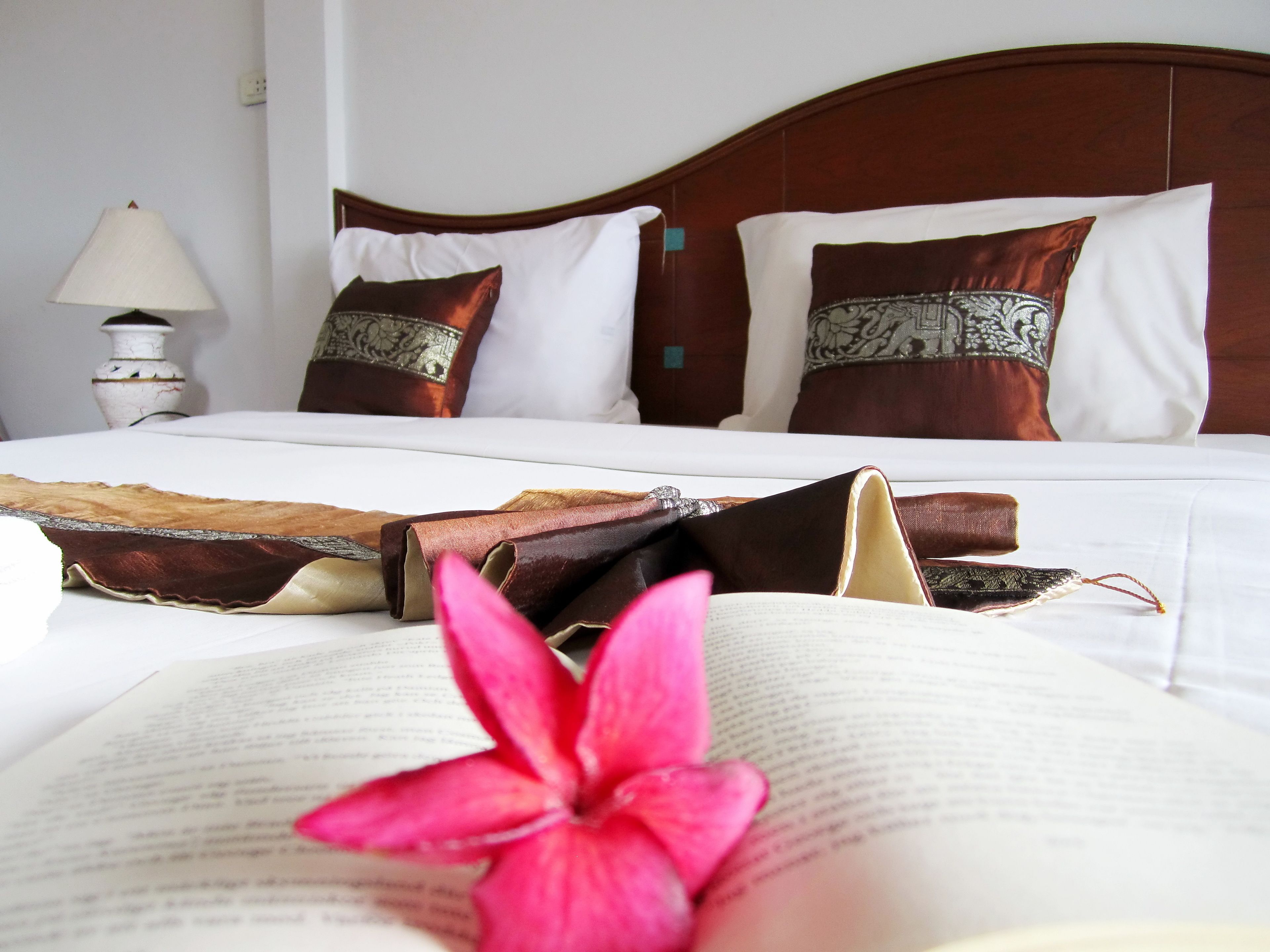 Una cama bien hecha con sábanas blancas y almohadas decorativas, una lámpara de noche y un libro abierto con una flor rosa encima.