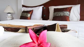 Una cama bien hecha con sábanas blancas y almohadas decorativas, una lámpara de noche y un libro abierto con una flor rosa encima.