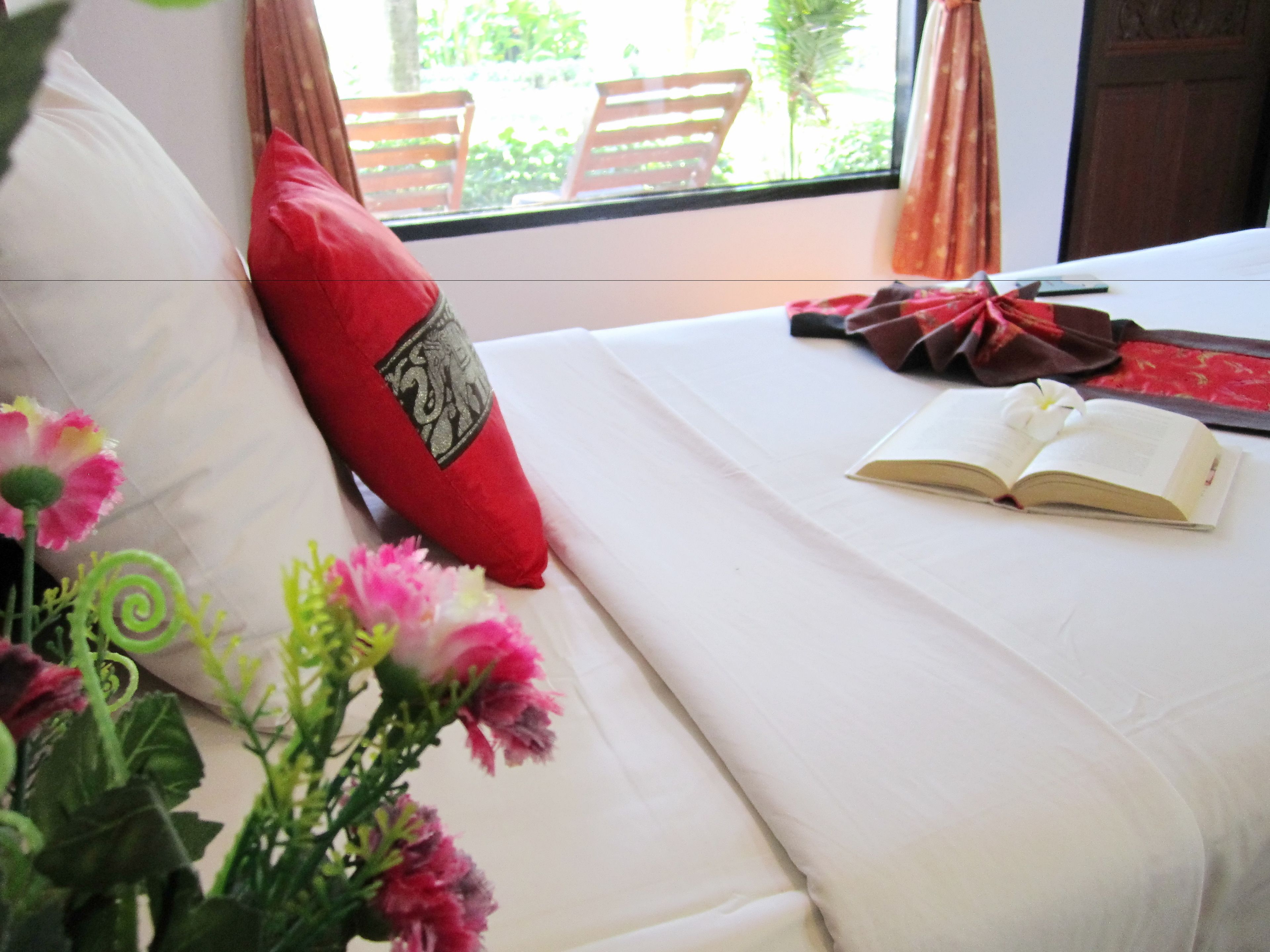 Una cama bien hecha con un almohadón rojo, un libro abierto y un ramo de flores.