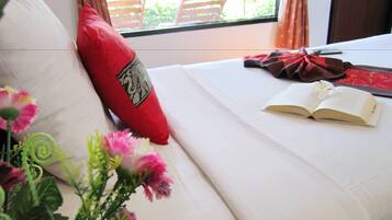 Una cama bien hecha con un almohadón rojo, un libro abierto y un ramo de flores.