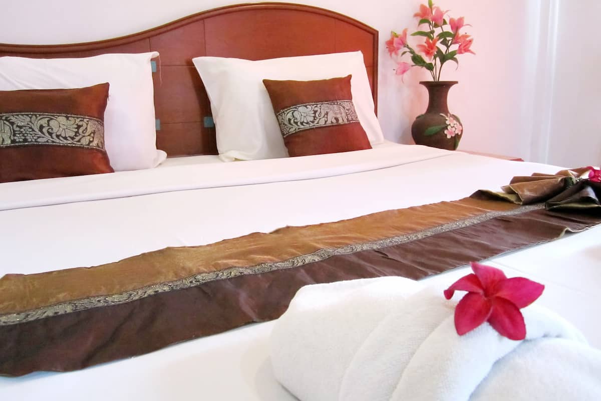 Una cama bien hecha con una colcha marrón y blanca, dos almohadas decorativas y un jarrón con flores rosadas sobre una mesita de noche.