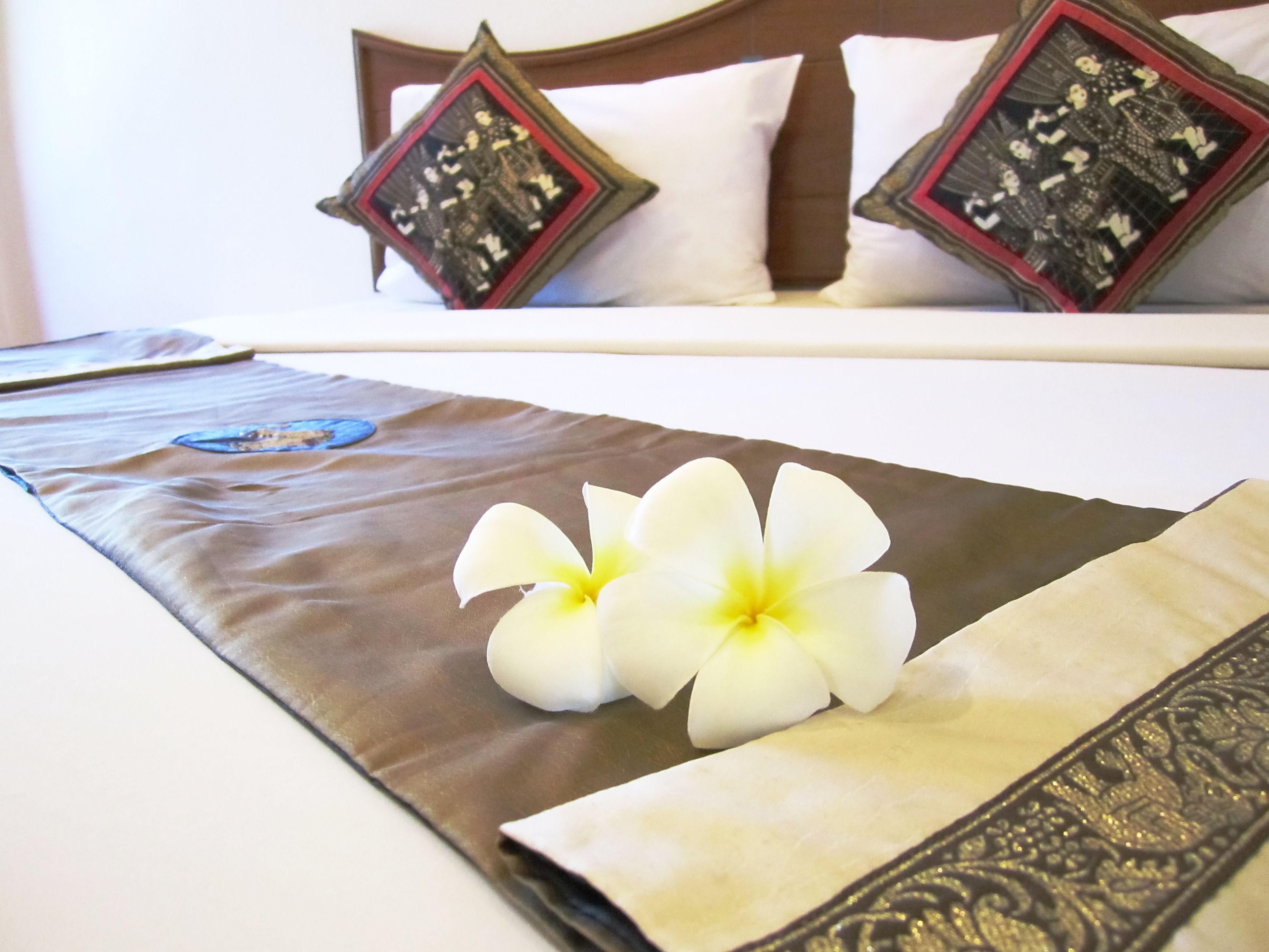 A neatly made bed with white linens, two decorative pillows with intricate designs, and a brown bedspread with a floral arrangement.