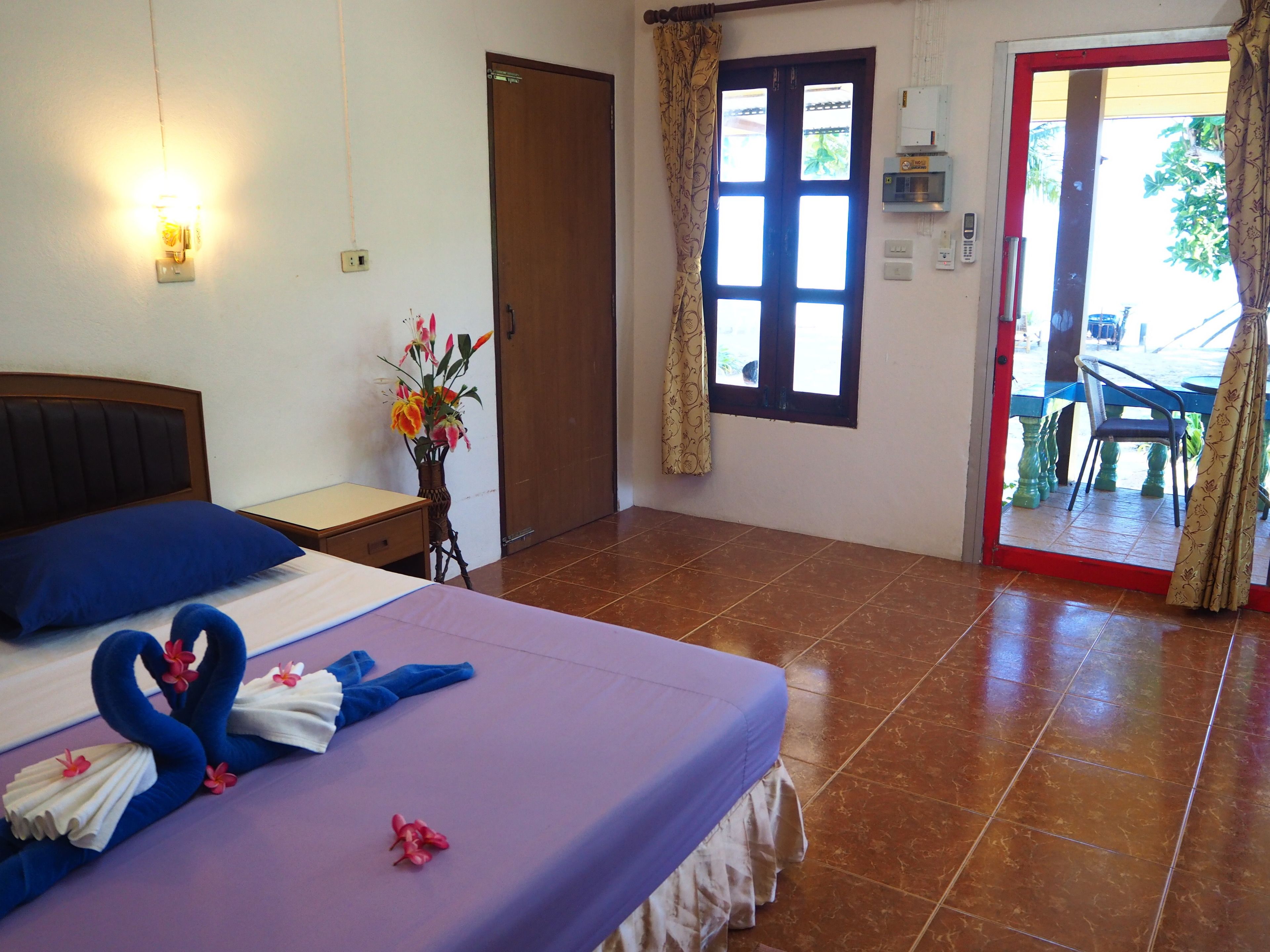 A hotel room with a bed, a nightstand, a vase with flowers, a window with curtains, and a view of the outside.