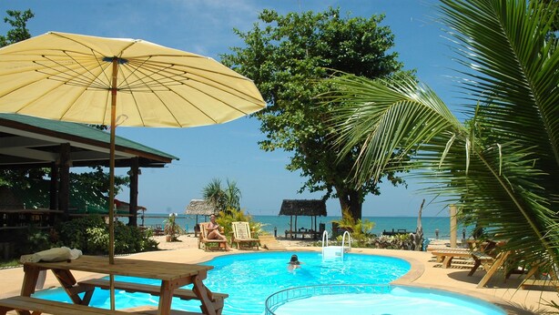 Un espace piscine avec une table et des bancs en bois, un parasol jaune et une vue sur l’océan.