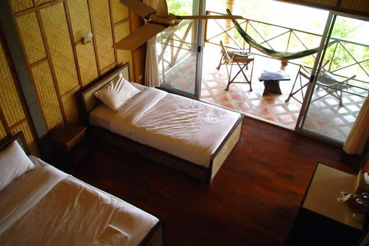 A room with two beds, a wooden floor, a window with a view of outdoor furniture, and a small table with a lamp.