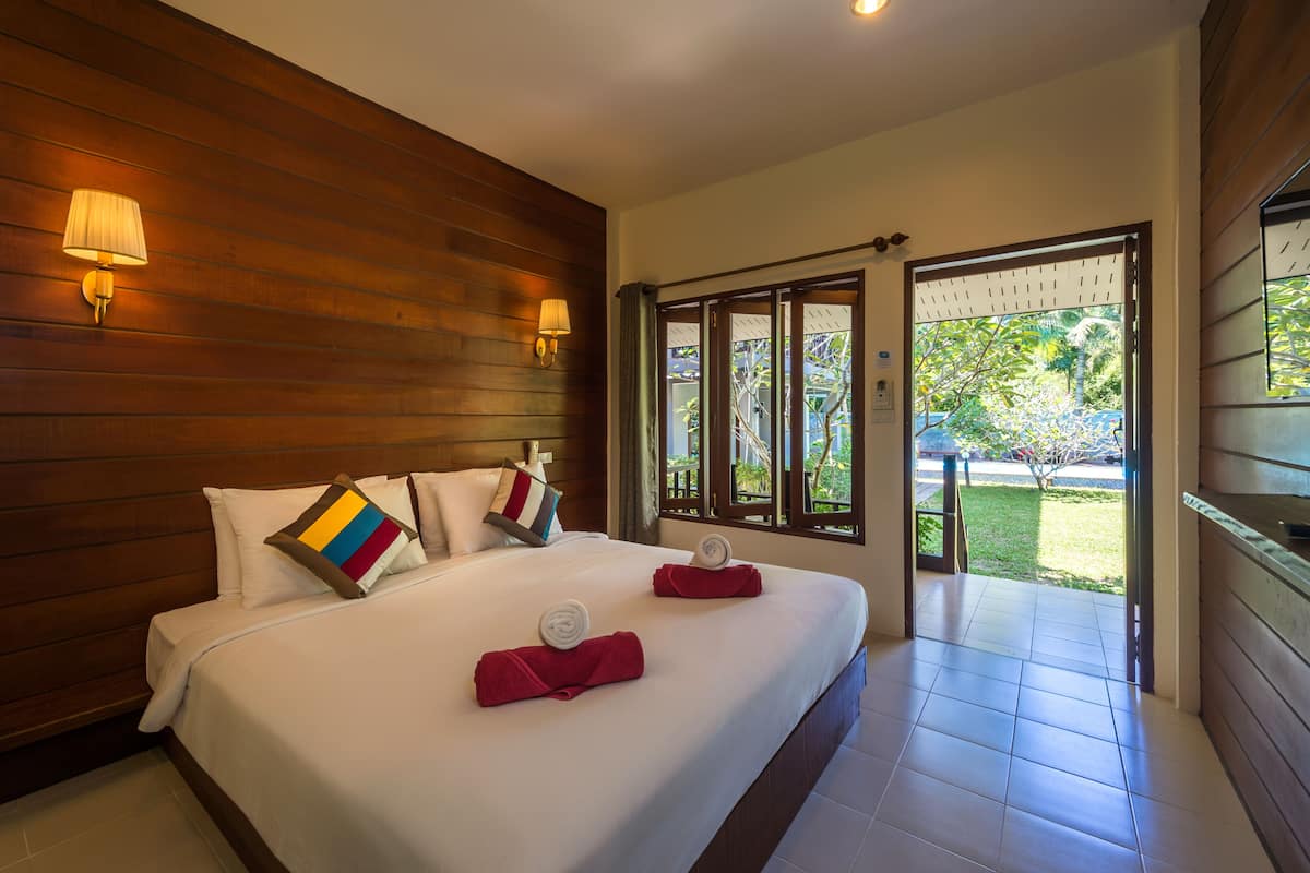 Habitación de hotel con cabecera de madera, cama grande con ropa blanca, dos lámparas de noche y vista a un jardín a través de un ventanal.