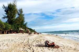 Una playa con una pareja relajándose en una tumbona, rodeada de palmeras y cabañas de playa.