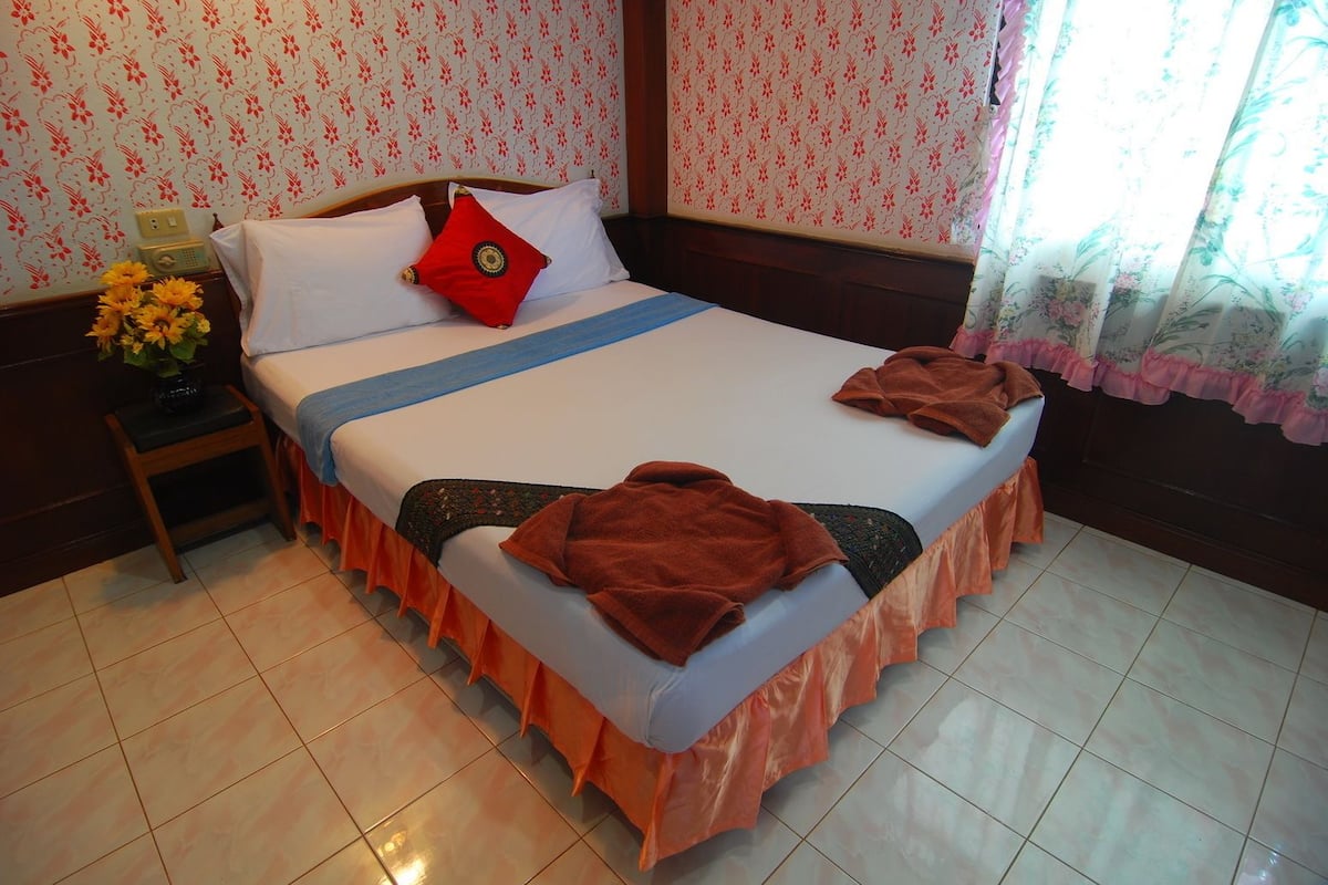 A neatly made bed with a red pillow, a blue blanket, and two brown towels on the side. There is a small wooden nightstand with a vase of yellow flowers and a radio beside the bed. The room has floral wallpaper and a window with curtains.