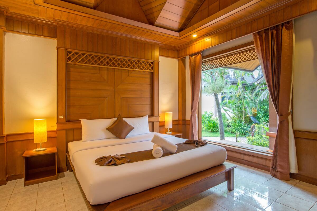 A spacious bedroom with a large bed, wooden headboard, bedside lamps, and a view of greenery outside.