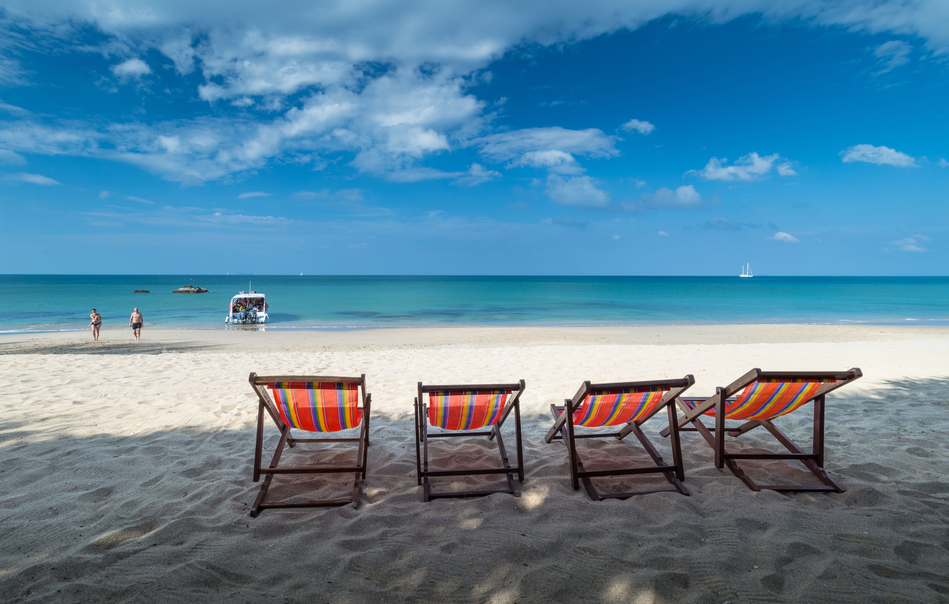 A beach with three wooden lounge chairs with striped cushions, a sandy beach, and a clear blue sky.