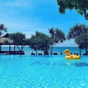Piscina azul cristalina con un pato inflable amarillo, rodeada de palmeras y una terraza de madera con sillones de descanso.