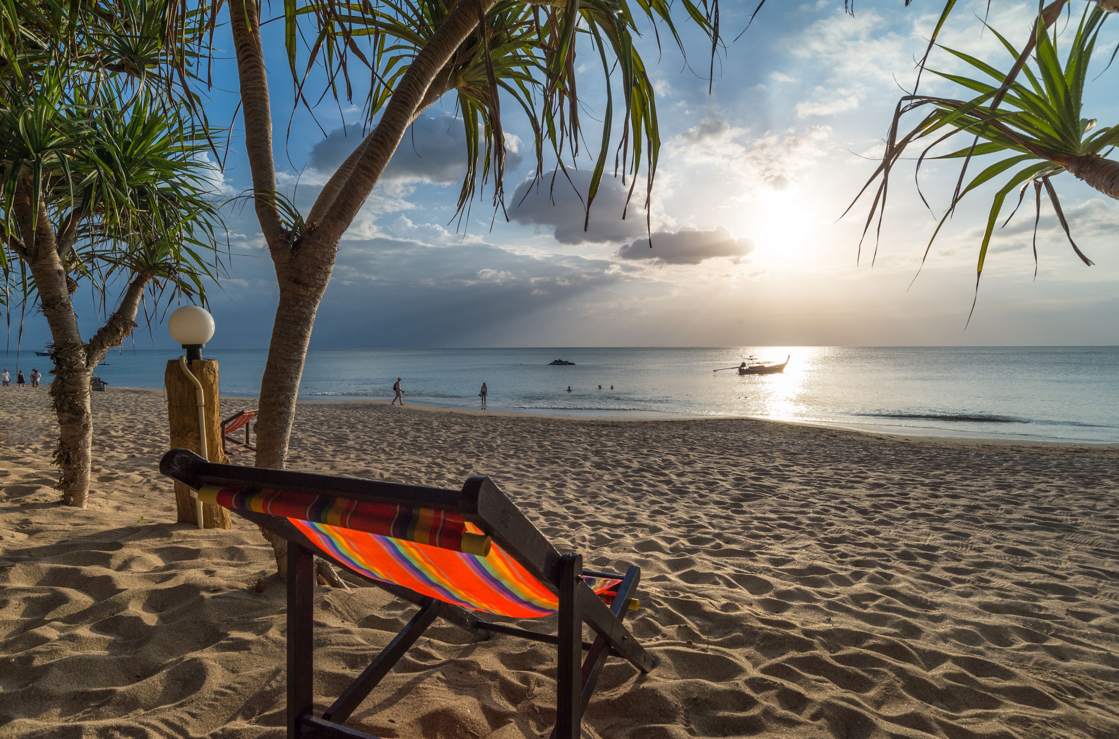 Uma praia com uma cadeira de praia colorida, palmeiras e um barco na água.