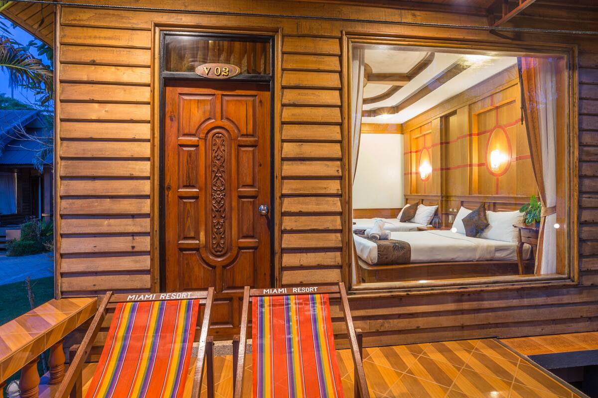 A wooden cabin with a large window, a door with a number plate, and a colorful striped bench outside.