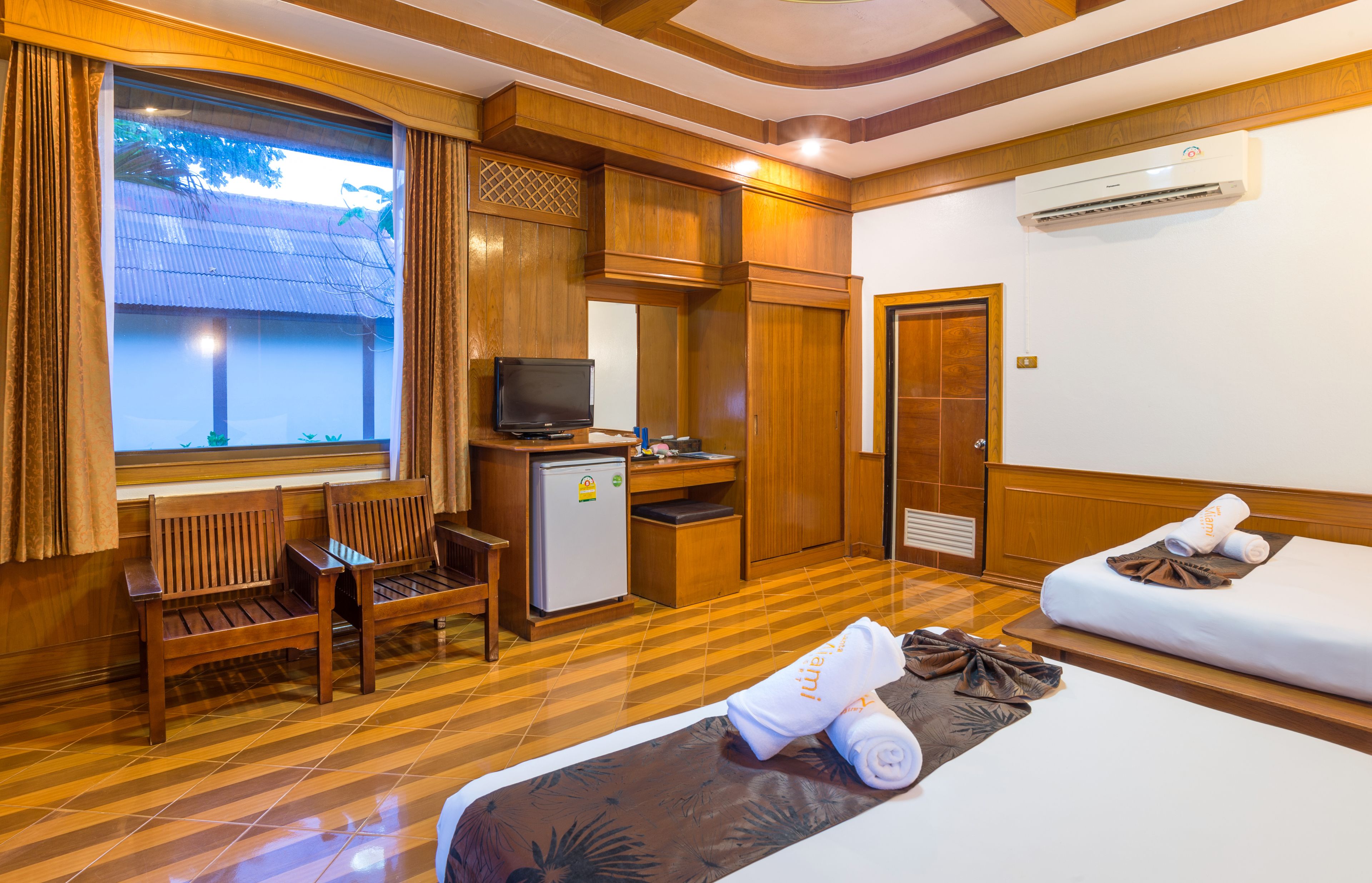 A hotel room with a wooden floor, a desk with a television, a mini-fridge, a bed with towels, and a window with curtains.