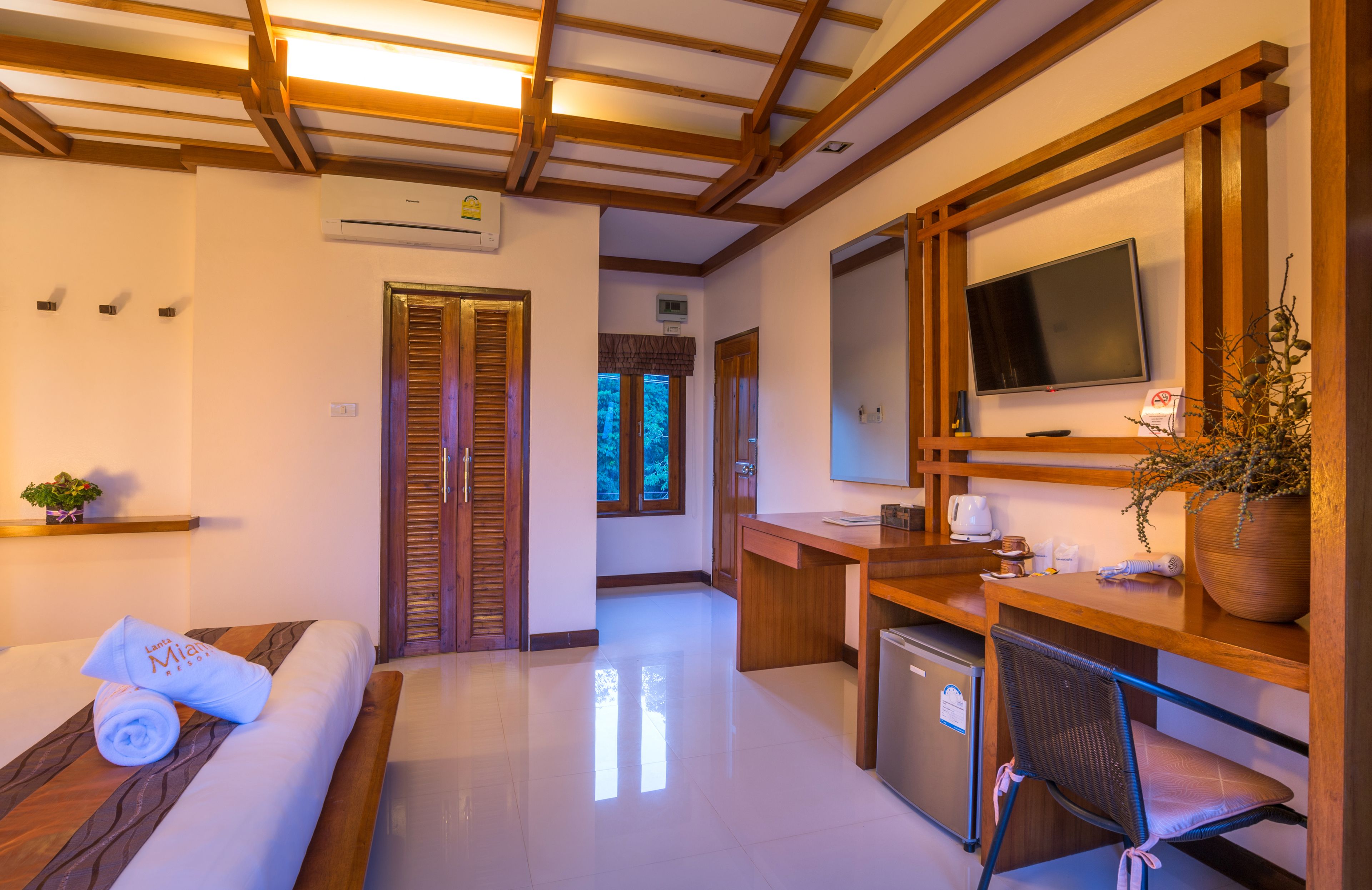 A hotel room with a wooden ceiling, a bed with a rolled towel, a desk with a computer, a TV, and a small kitchenette.
