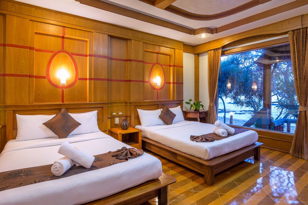 A hotel room with two beds, wooden paneling, and a large window with a view of palm trees.