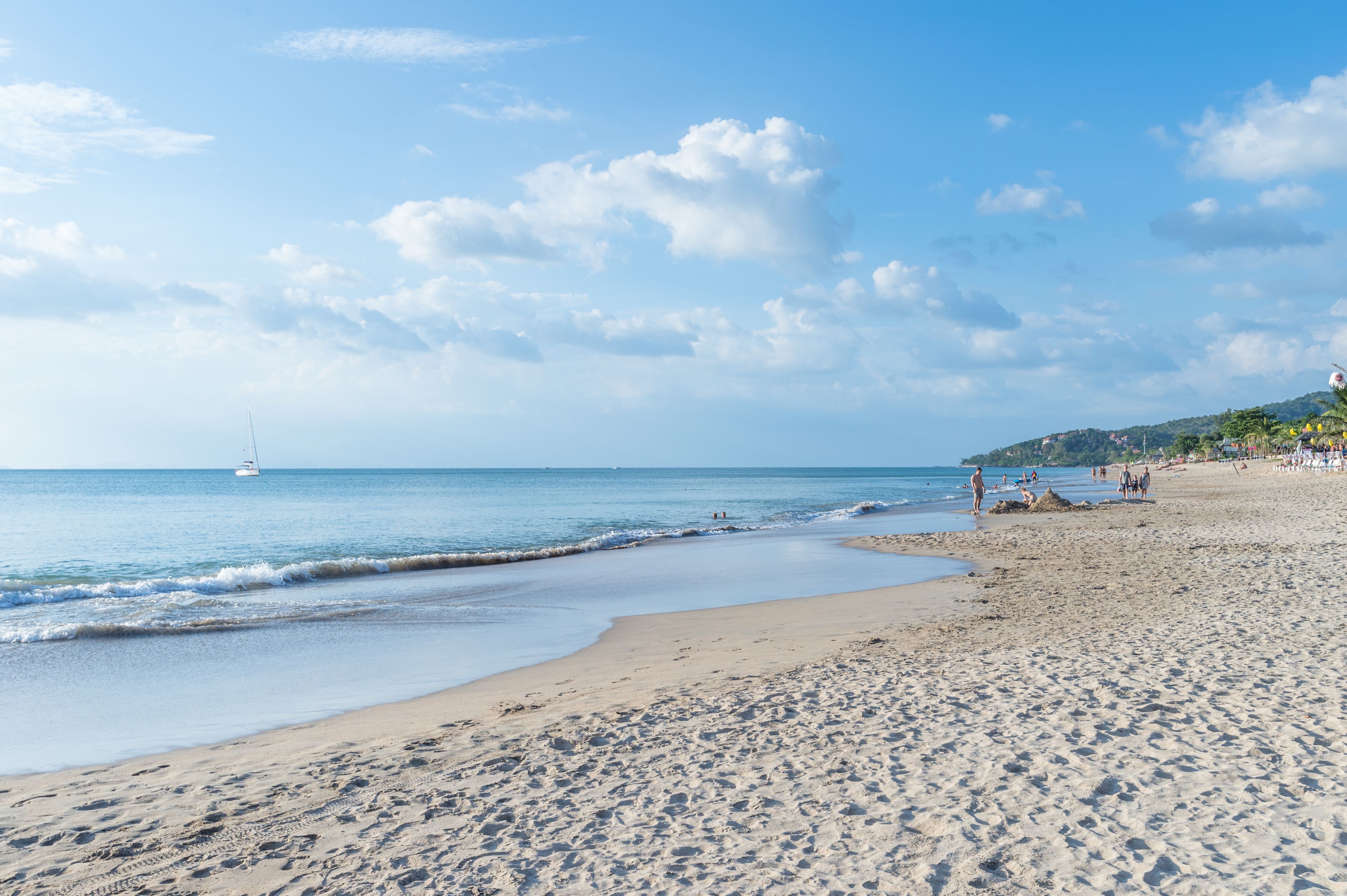 A beach with a sailboat in the distance, sandy shore, and people walking.