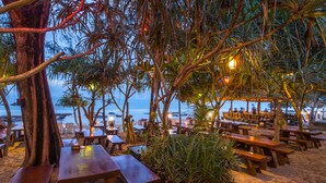 Un restaurante frente a la playa con mesas y bancos de madera, rodeado de palmeras y vistas al océano.