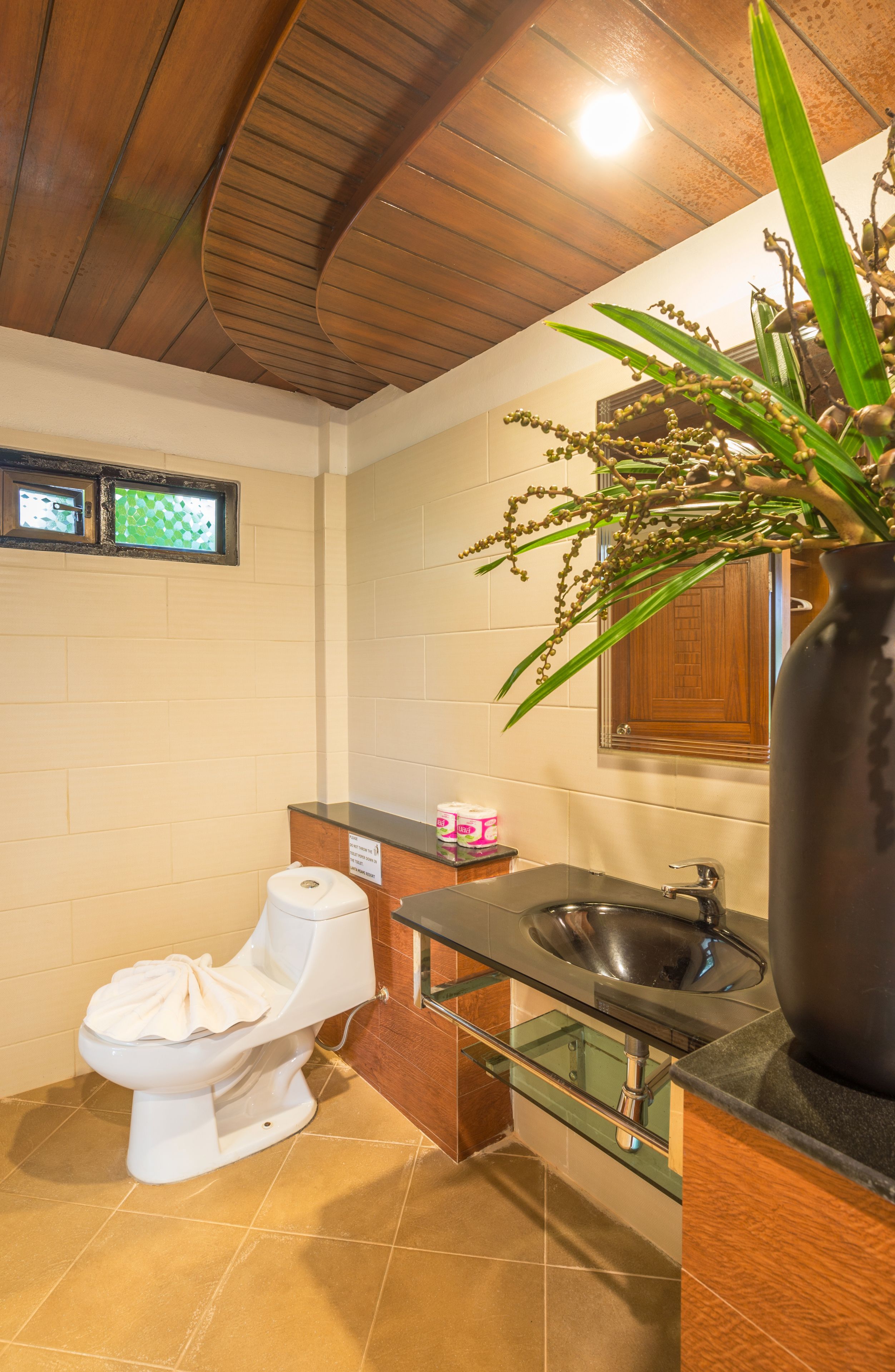 Un bagno con soffitto in legno, un water bianco, un lavandino in vetro e una grande pianta in vaso.