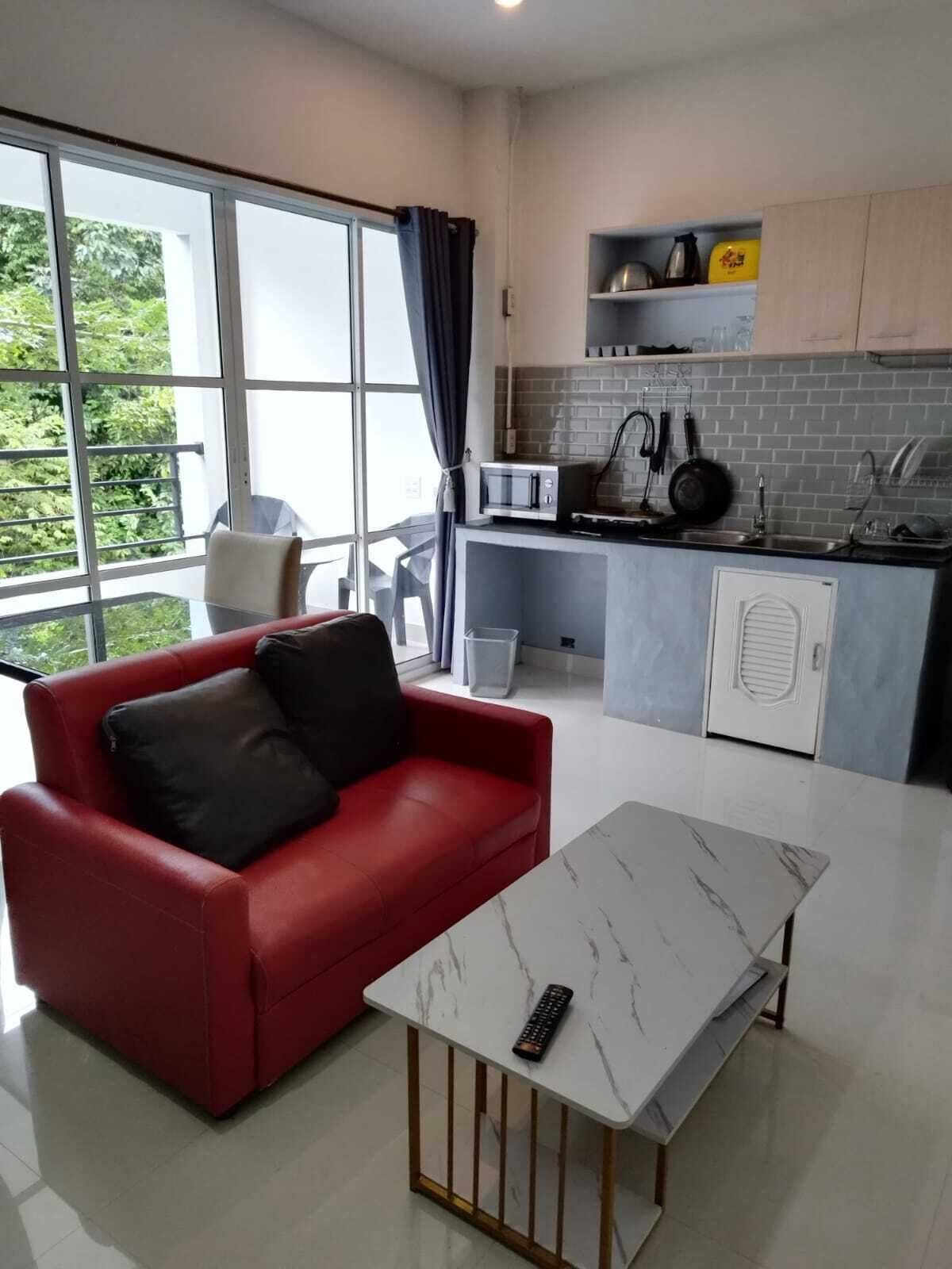 A modern living room with a red sofa and a white table, a kitchen with grey cabinets and a microwave, and large windows with curtains.