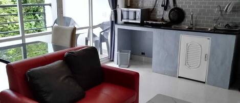 A modern living room with a red sofa and a white table, a kitchen with grey cabinets and a microwave, and large windows with curtains.