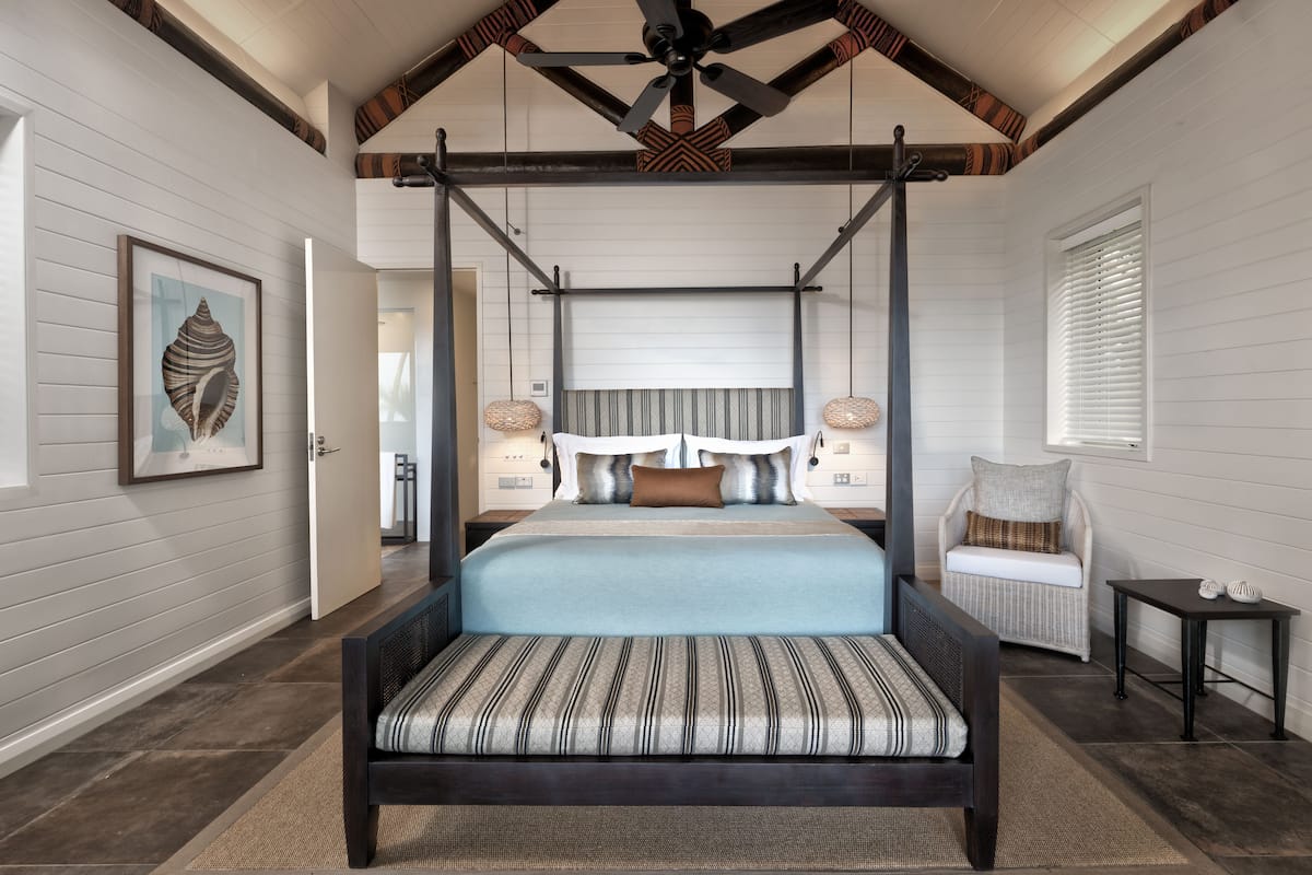 A bedroom with a four-poster bed, a large framed artwork on the wall, and a wicker chair by the window.