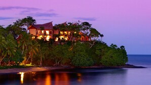 A large, multi-story house with a thatched roof, surrounded by lush greenery and overlooking a calm body of water. - Nanuku Resort Fiji (Pacific Harbour)