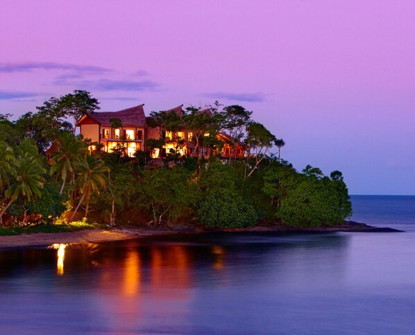 A large, multi-story house with a thatched roof, surrounded by lush greenery and overlooking a calm body of water. - Nanuku Resort Fiji (Pacific Harbour)