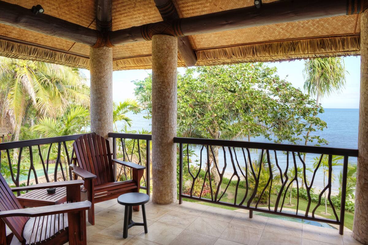 A covered outdoor seating area with chairs and a table, overlooking a beach and palm trees.