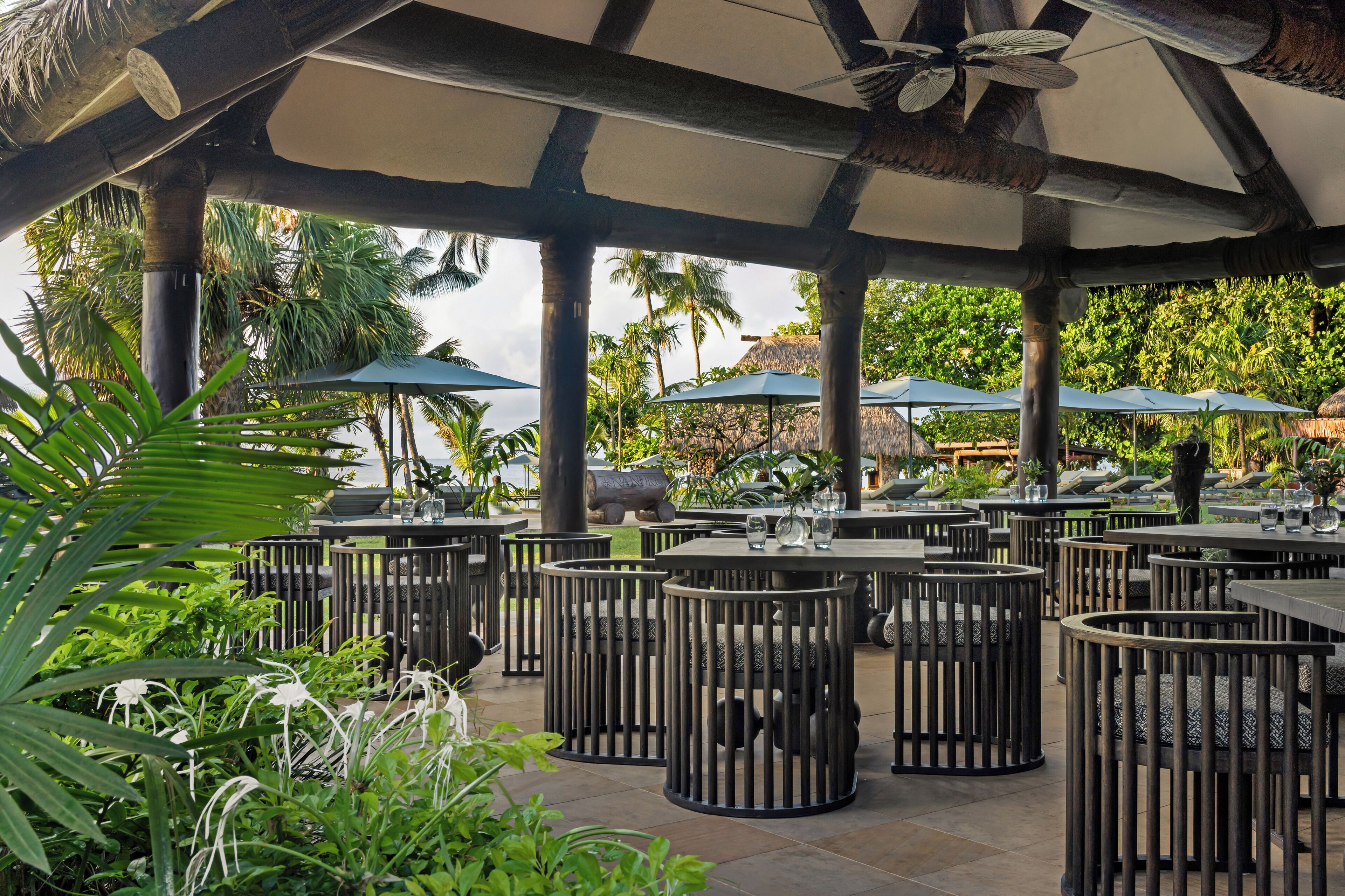 Un espace extérieur aménagé en terrasse, avec des tables et des chaises rondes, entouré de palmiers et agrémenté de parasols.