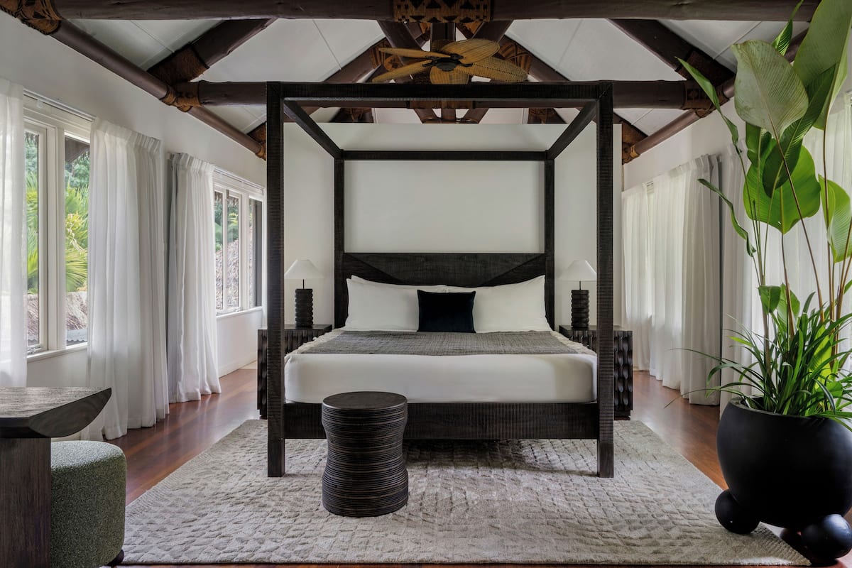 A bedroom with a four-poster bed, a round side table, a potted plant, and a view of greenery outside.