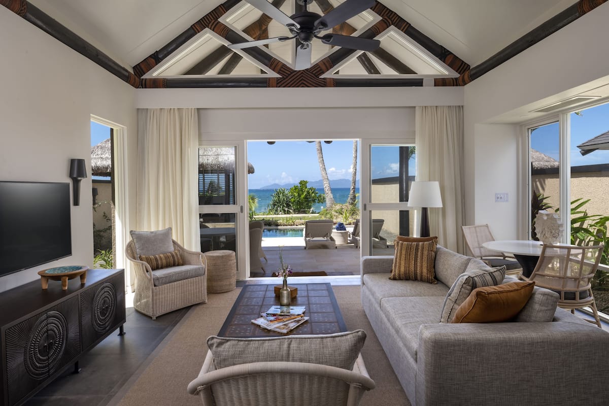 A spacious living room with a large TV, comfortable seating, a coffee table, and a view of the ocean through large windows.