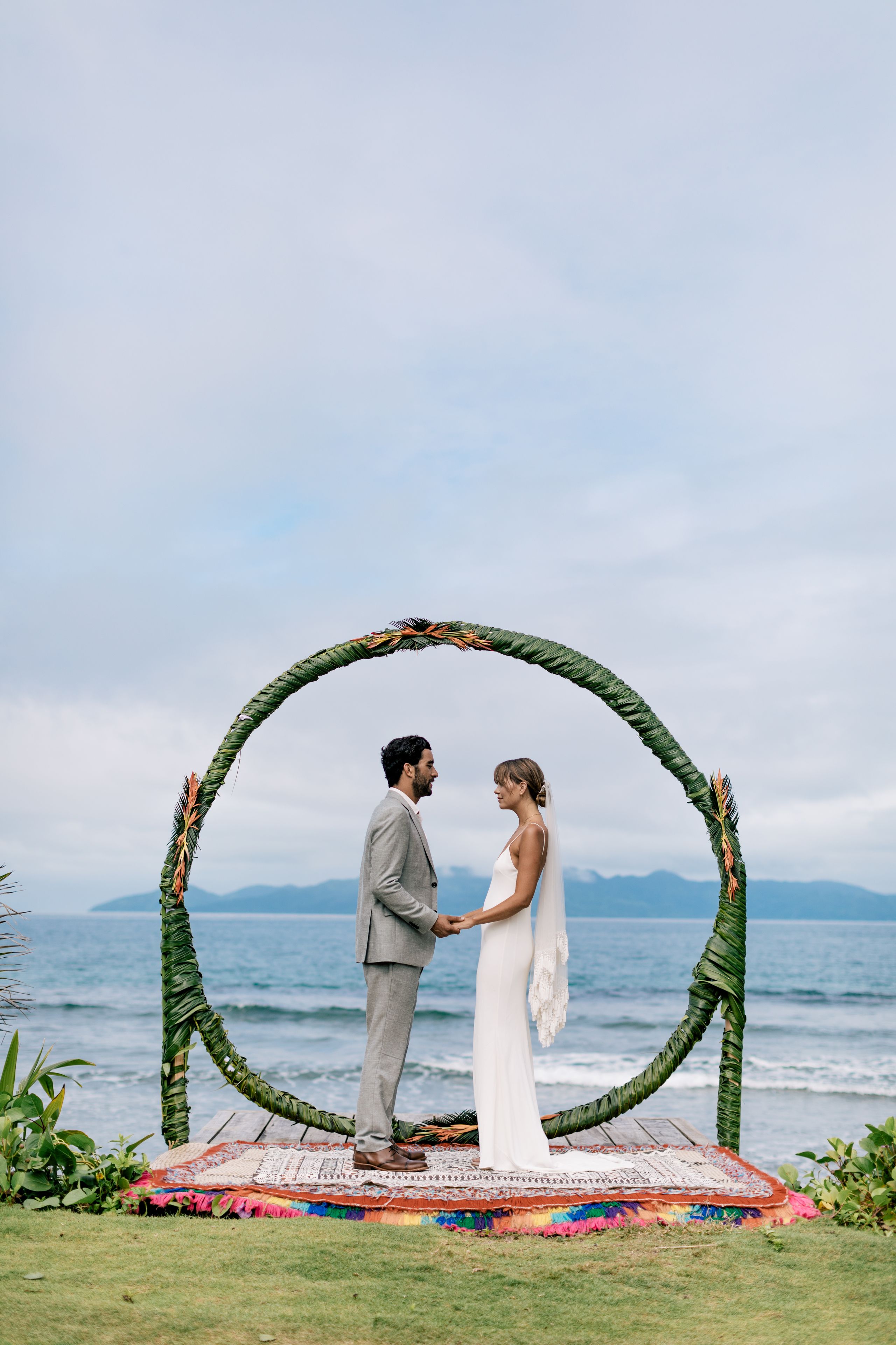 A couple standing under a circular arch decorated with foliage, holding hands, with a scenic ocean view in the background.