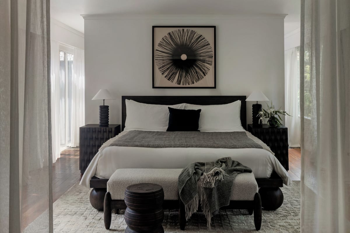 A modern bedroom with a large bed, two bedside tables with lamps, a wall art piece, and a view of the outdoors.