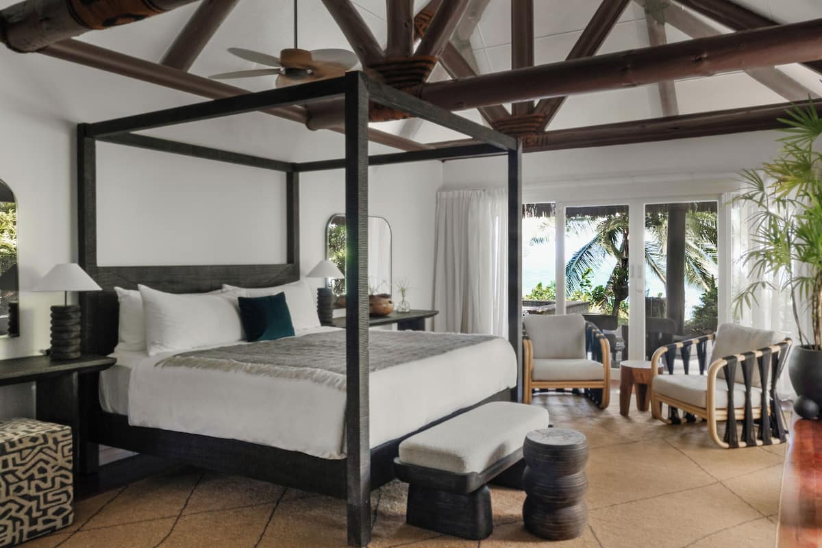 A four-poster bed with a headboard, bedside tables, a bench, and a chair in a room with a tiled floor and a view of palm trees.