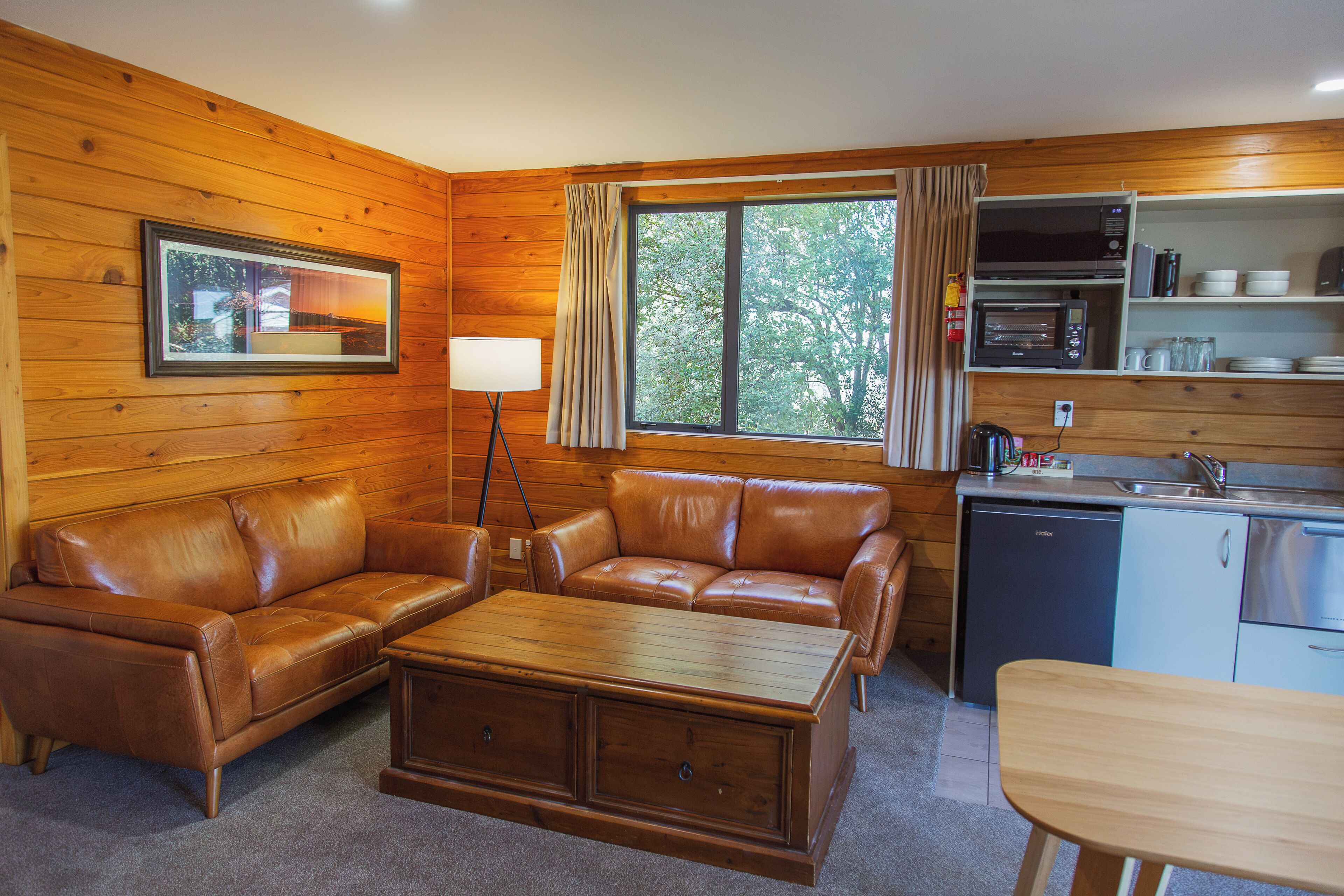 Interior acogedor de una cabaña con detalles de madera, un sofá, una mesa de centro, una mesa de comedor, una zona de cocina con microondas y fregadero, y una ventana con cortinas.