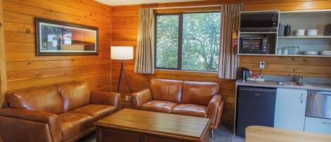 Interior acogedor de una cabaña con detalles de madera, un sofá, una mesa de centro, una mesa de comedor, una zona de cocina con microondas y fregadero, y una ventana con cortinas.