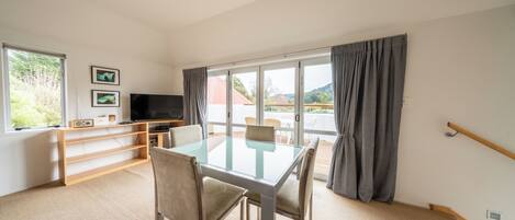 A modern room with a glass-top table, four chairs, a flat-screen TV, a bookshelf, and a large window with curtains.