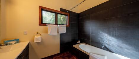 A modern bathroom with a wooden floor, a white bathtub, and a window with a view of greenery.