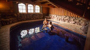 Una piscina interior con paredes de piedra, suelo de baldosas, bancos de madera y un muro de piedra con una ventana.