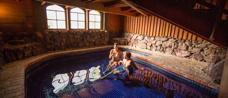 A stone-walled indoor pool with a tiled floor, wooden benches, and a stone wall with a window.