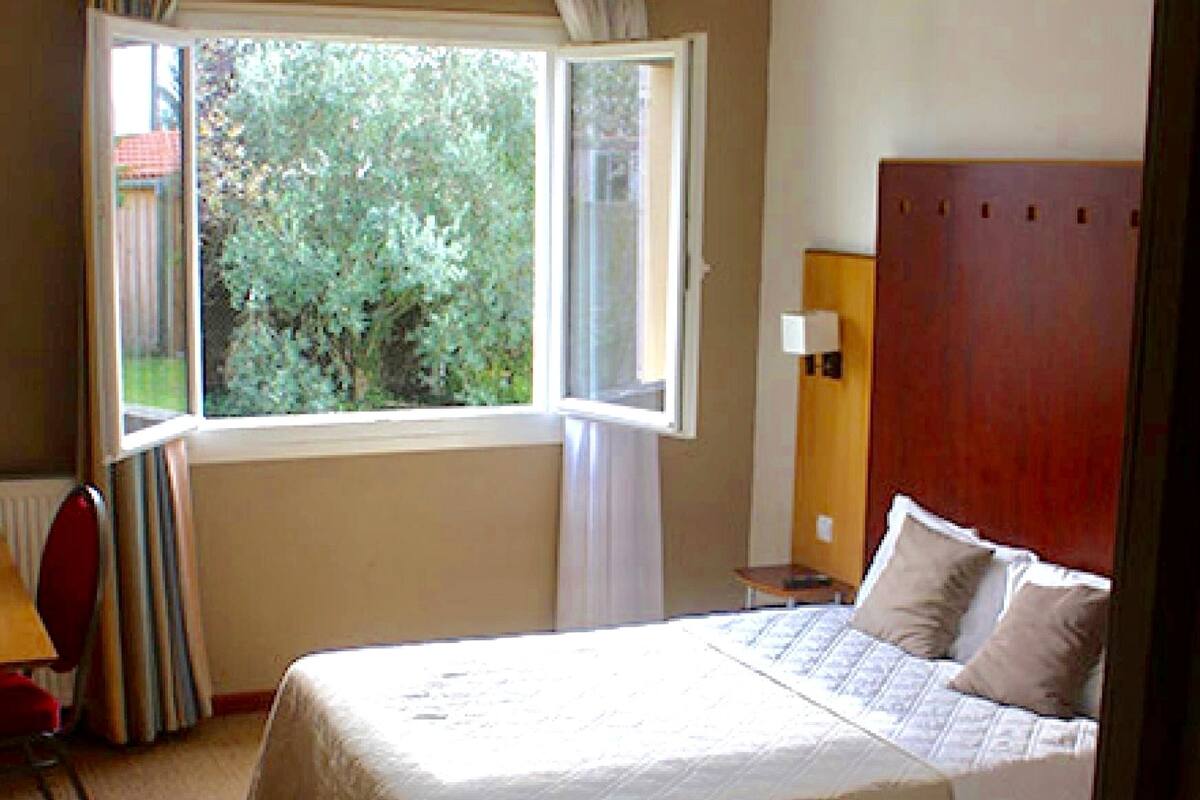 A hotel room with a bed, a window with curtains, a chair, a bedside table, and a lamp.
